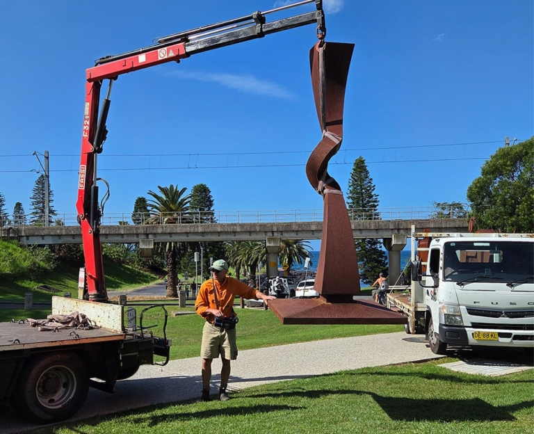 Tall Corten steel sculpture being installed by truck crane in a public park