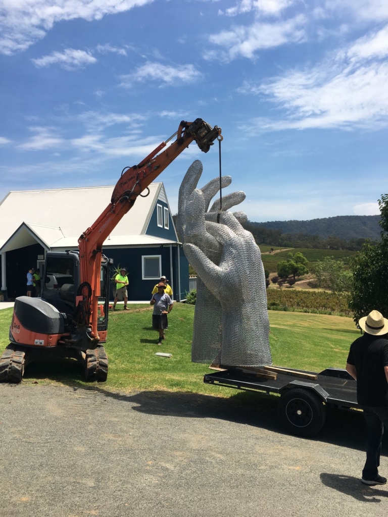 Large hand sculpture being positioned by excavator on a lawn